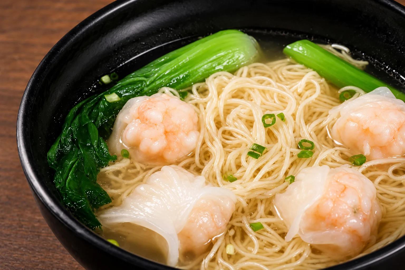 Black bowl of thin noodles in clear broth with shrimp dumplings, bok choy and spring onion.